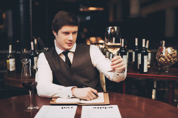 Confident sommelier makes wine card in restaurant, making notes and viewing glass of white wine. - Foto, Bild