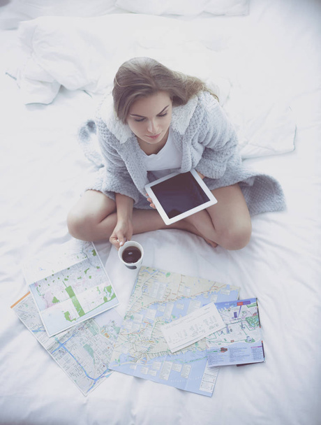 Relaxed young woman sitting on bed with a cup of coffee and digital tablet - Foto, afbeelding