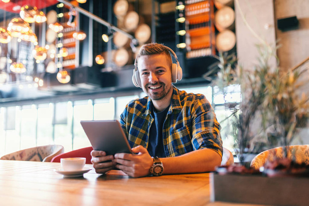 Smiling freelancer looking at camera, having headphones on ears and tablet in hands. In front of him on a table coffee. Cafeteria interior. - Photo, Image