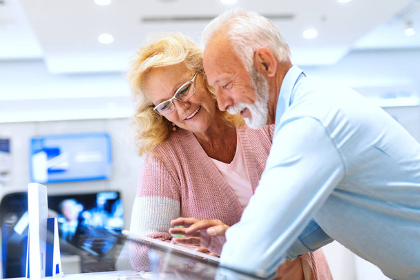 Sonriendo Caucásico senior pareja vestida casual prueba tableta mientras se apoya en el soporte en la tienda de tecnología
. - Foto, imagen