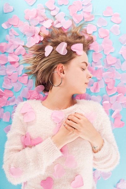 top view of beautiful girl with eyes closed lying with heart shaped confetti isolated on blue, st valentines day concept - Zdjęcie, obraz