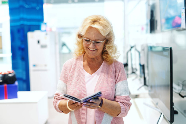 Smiling old blonde Caucasian woman with eyeglasses and dressed casual looking at brochure while standing in tech store. - Fotografie, Obrázek