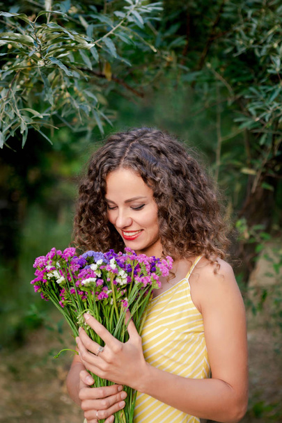Молода жінка з барвистими квітами в руках. Приваблива усміхнена жінка з кучерявим коричневим волоссям в літньому парку. Концепція жіночого дня
. - Фото, зображення