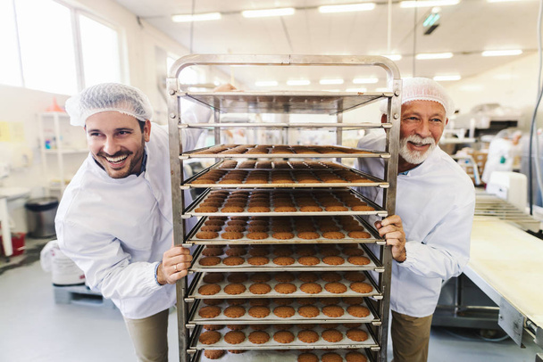 Deux employés souriants qui travaillent dur en uniforme blanc stérile poussant étagère avec des biscuits. Usine alimentaire intérieur
. - Photo, image