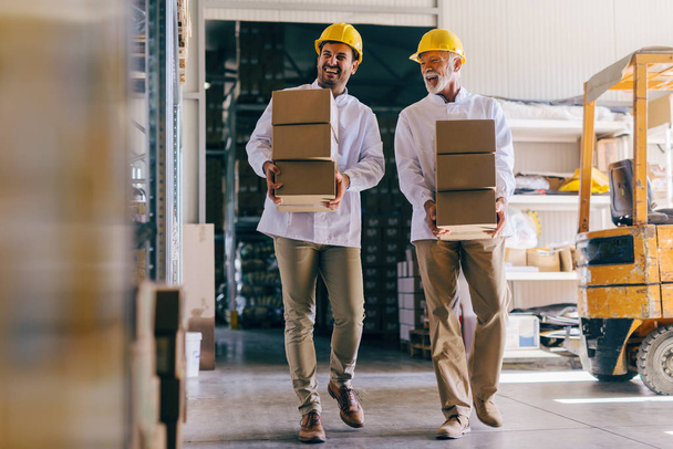 Двое улыбающихся рабочих в белых мундирах и в шлемах на головах несут коробки на складе
. - Фото, изображение