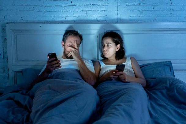 Couple affected by technology addiction ignoring at each other in apathy and anger lying in bed using their smart phones late at night in darkness in relationship issues and internet mobile addiction. - 写真・画像
