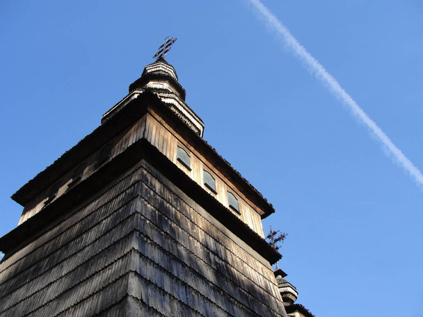 Antigua iglesia ortodoxa de madera, edificio con techo de tejas de madera. Huella blanca desde el avión en el fondo del cielo azul. Avión sale detrás de una racha blanca a través del cielo
 - Foto, Imagen