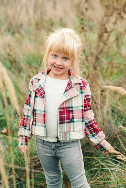 Menina bonito andando no campo. Menina elegante com olhos azuis e cabelo loiro. Moda infantil. Criança feliz no pôr do sol de outono na natureza
 - Foto, Imagem