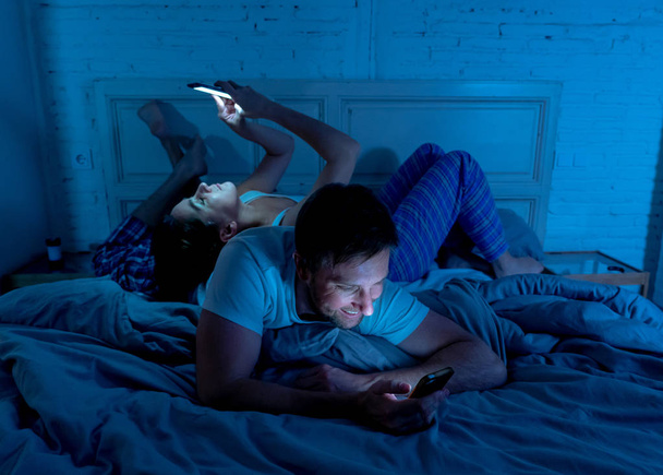 Sad man and woman married couple using their smart mobile phone in bed at night ignoring each other as strangers in relationship and communication problems and internet social network addiction. - Foto, Imagen
