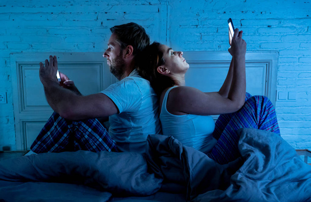 Sad man and woman married couple using their smart mobile phone in bed at night ignoring each other as strangers in relationship and communication problems and internet social network addiction. - Foto, Bild