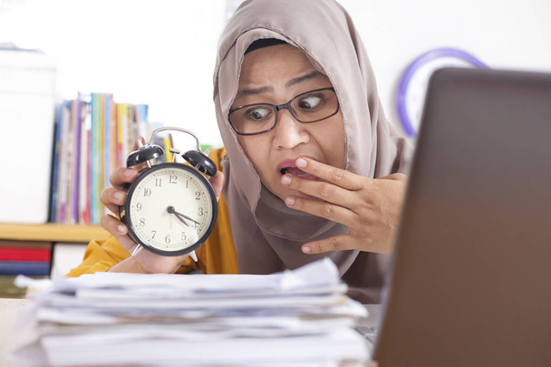 Young Asian businesswoman wearing suit and hijab worried by time when looking at a clock. Work deadline stress at the office - Fotografie, Obrázek
