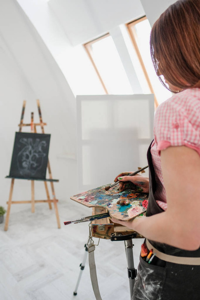 Schilder van het mooie meisje in een witte studio tekent op een ezel op canvas. - Foto, afbeelding