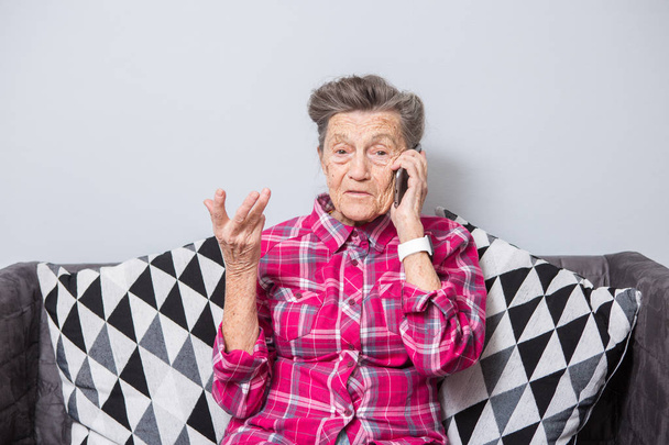 Thème personne âgée utilise la technologie. Mature joie heureuse sourire active cheveux gris rides caucasiennes femme assise à la maison dans le salon sur le canapé et à l'aide d'un téléphone mobile, appelant et parlant au téléphone
. - Photo, image