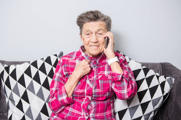 Thema alter Mensch nutzt Technologie. Ältere glückliche Freude Lächeln aktive graue Haare kaukasische Falten Frau sitzt zu Hause im Wohnzimmer auf dem Sofa und mit einem Handy, telefonieren und telefonieren. - Foto, Bild