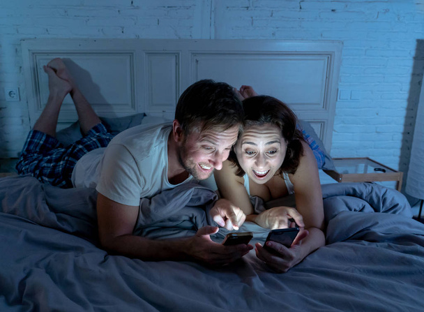 Smiling attractive young men and woman couple using smartphone lying on bed together. Happy boyfriend and girlfriend checking social networks or mobile app in gaming and technology addiction concept. - Photo, image