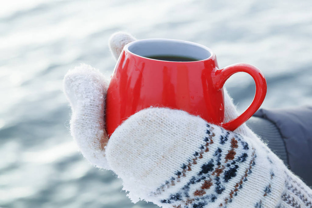 Hands in woolen mittens hold a red mug with hot tea or coffee, standing on the snow. Picnic on a frosty Sunny day. The concept of warming on a winter walk - Foto, afbeelding