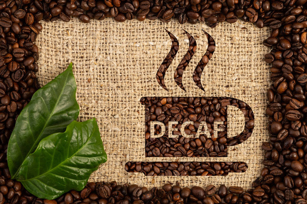 Cup with decaf text written as aroma of no-caffeine hot beverage inside scattered coffee beans frame on brown burlap bag background - Photo, Image