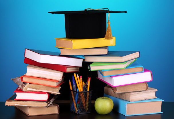Books and magister cap against school board on wooden table on blue background - Foto, afbeelding