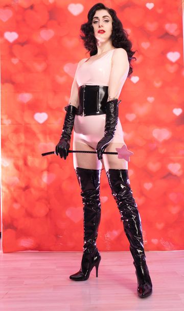 pinup brunette woman wearing pink latex rubber costume with black fetish corset and overknee boots standing with bdsm riding crop on red background with hearts - Photo, Image