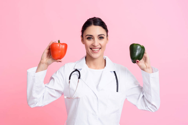 Portrait of smiling female doctor with stethoscope holding red and green peppers isolated. - Foto, imagen
