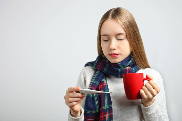 Chory dziewczynka kaukaski w szalik z kubek gorącej herbaty i Termometr elektroniczny na neutralnym tle. Koncepcja zdrowia, ciepła gorączka, wirus. - Zdjęcie, obraz