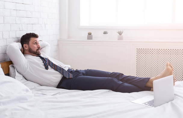 Liikemies rentouttava työskentelemästä kannettavan, makaa sängyssä
 - Valokuva, kuva