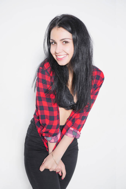 Thoughtful young woman posing near white sofa, dressed in plaid shirt and black pants - Photo, Image