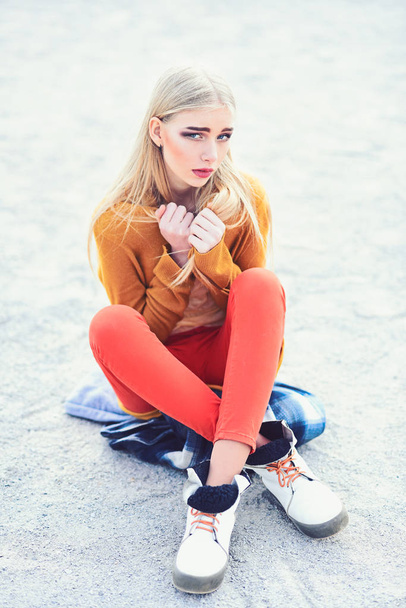 Young blond model with straight hair in bright outfit sitting on street with legs crossed and holding her arms crossed at her chest - Foto, imagen