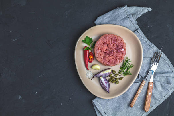 Delicious steak tartare and ingredients on ceramic plate, set of cutlery knife, fork on black stone concrete textured surface background. Copy space background, top view flat lay. - Foto, Imagem