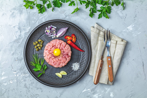 Steak tartare with yolk and ingredients on black ceramic plate, set of cutlery knife and fork on light gray stone concrete textured surface background. Copy space background, top view flat lay. - Photo, Image