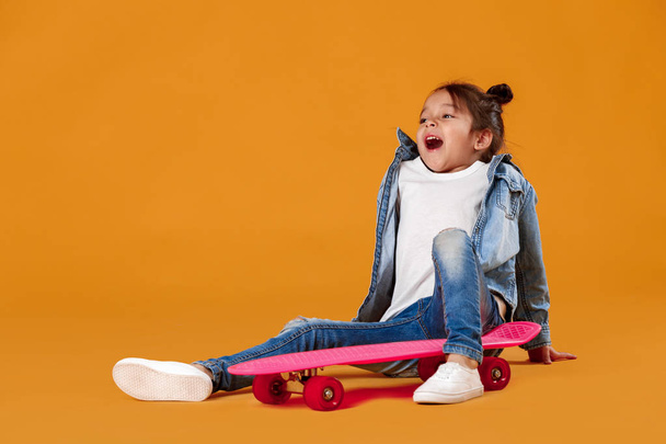 Stylish little child girl with skateboard in denim on orange background - Fotoğraf, Görsel