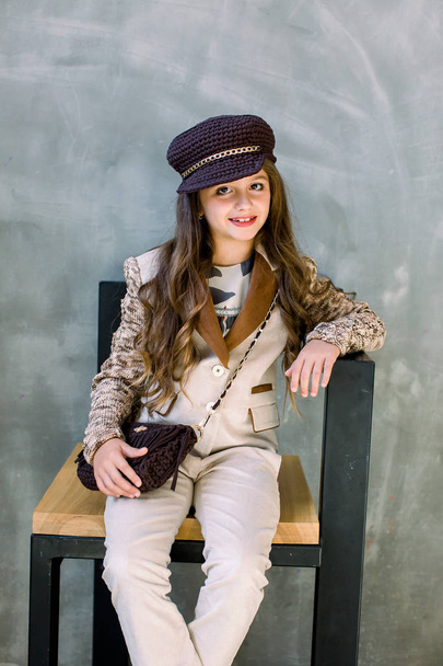 Portrait of adorable smiling little girl in a light suit, a golden sneakers and a hat isolated on a grey sitting on the chair - Foto, afbeelding