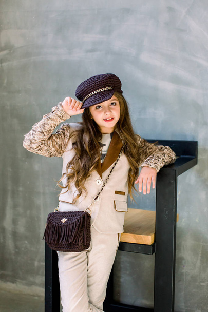 Portrait of cute little girl in a light suit, a golden sneakers and a hat on a gray background stands next to a wooden chair - 写真・画像