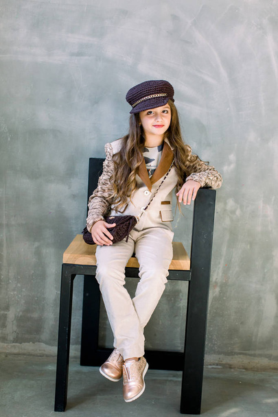 Portrait of adorable smiling little girl in a light suit, a golden sneakers and a hat isolated on a grey sitting on the chair - Foto, immagini