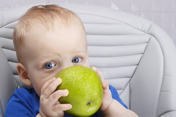 niedliches Baby 1,4 Jahre alt sitzt auf hohem Kinderstuhl und isst grünen Apfel allein in weißer Küche - Foto, Bild