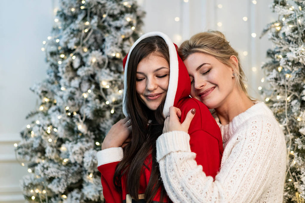 zwei schöne Schwestern in Pullovern, die sich in der Nähe des Weihnachtsbaums umarmen - Foto, Bild