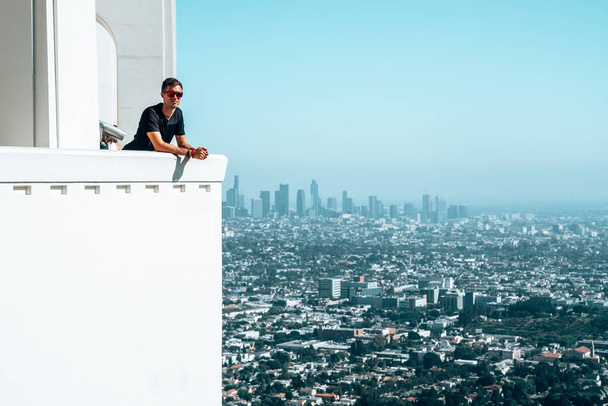 Νεαρός άνδρας κάθεται με θέα στο κέντρο της πόλης Λος Άντζελες στο παρασκήνιο. Καταπληκτικό Χόλιγουντ. - Φωτογραφία, εικόνα