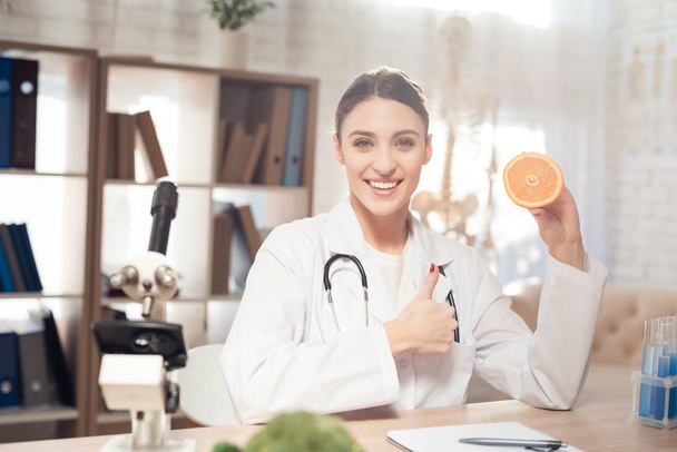 Female doctor sitting at desk in office with microscope and stethoscope. Woman is holding orange. - Photo, Image