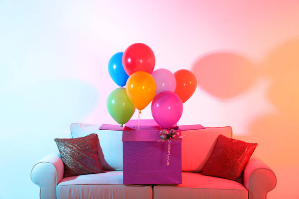 Gift box with bright air balloons on sofa against color background - Zdjęcie, obraz
