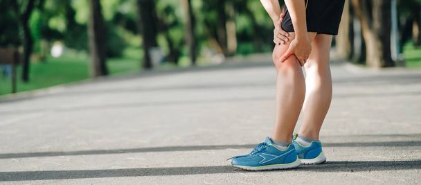 Young fitness woman holding his sports leg injury, muscle painful during training. Asian runner having knee ache and problem after running and exercise outside in summer - Photo, Image