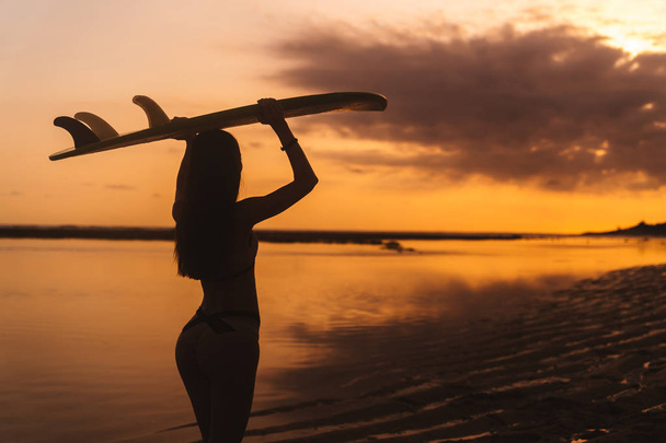 Silhouette eines schlanken Mädchens mit Surfbrett in den Händen am Strand vor dem Hintergrund des schönen Sonnenuntergangs - Foto, Bild