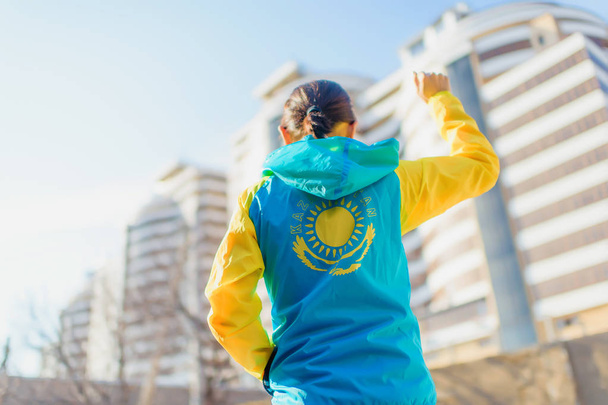 A man stands in a sports jacket on the background of buildings. The girl raised her hand with a clenched fist, holding the other hand in her pocket. Yellow and blue sports jacket. Yellow symbol of the sun and the eagle on a blue background. - Photo, Image