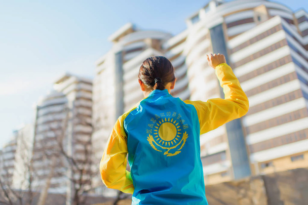 A man stands in a sports jacket on the background of buildings. The girl raised her hand with a clenched fist, holding the other hand in her pocket. Yellow and blue sports jacket. Yellow symbol of the sun and the eagle on a blue background. - Photo, Image