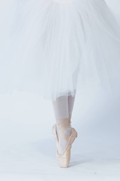 Ένα κορίτσι στέκεται στα πόδια του απλωμένα, παπουτσωμένος στα παπούτσια pointe. Ένα παιδί σε ένα λευκό φόρεμα για το χορό και λευκό καλσόν. Πόδια σε pointe. Ασκήσεις στη σχολή μπαλέτου. Μαθήματα μπαλέτου. Λευκό φόντο. - Φωτογραφία, εικόνα