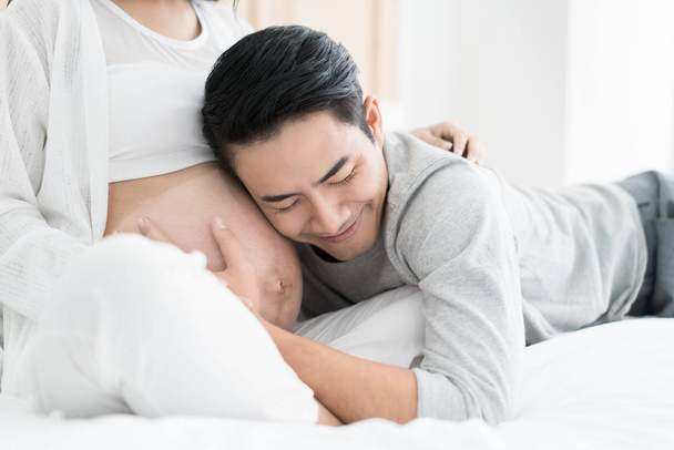 asiatischer gutaussehender Mann hört auf den Bauch seiner schönen schwangeren Frau und lächelt. Konzept der Familienliebe. - Foto, Bild
