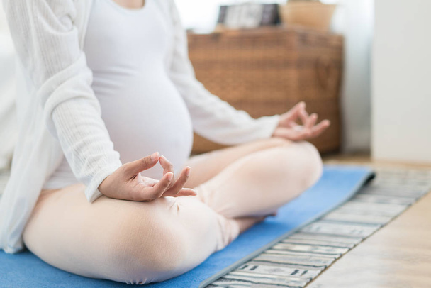 Meditating on maternity. Close-up of pregnant woman meditating while sitting in lotus position at home - Valokuva, kuva