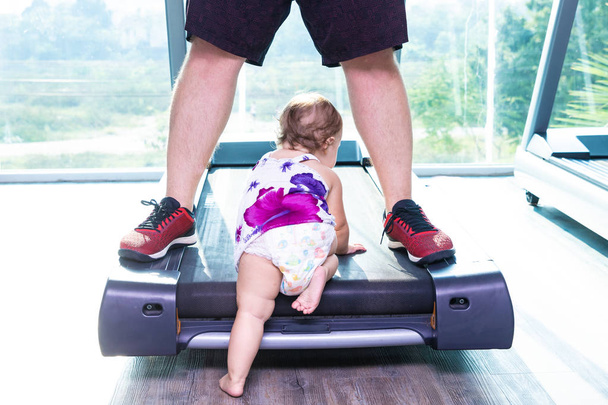 baby crawling on the treadmill in the gym with a view from the window on the coconut trees. Baby creeps between daddys legs - Fotografie, Obrázek