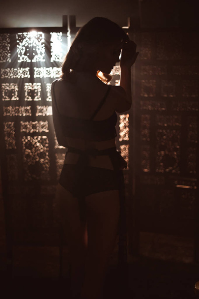 Silhouette of lovely young woman in a beautiful black old-fashion lace lingerie posing against a vintage wooden carved screen lit by beam of light. Attractive slender girl with long blond hair. - 写真・画像