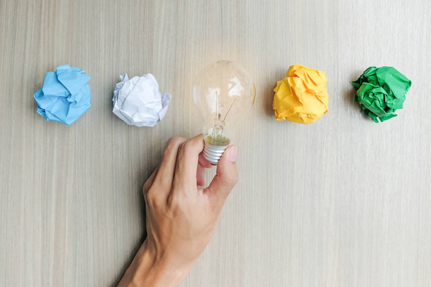 Mão segurando lâmpada ou lâmpada com papel colorido amassado na mesa de madeira. Novos conceitos de Ideia, Criatividade, Gênio e Inovação
 - Foto, Imagem