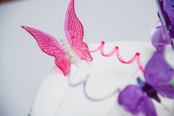 Белый торт украшен карамельными бабочками. Фиолетовые и розовые сладкие бабочки. Белый торт на белой тарелке. Украшение торта. Десерт. Кулинария
. - Фото, изображение
